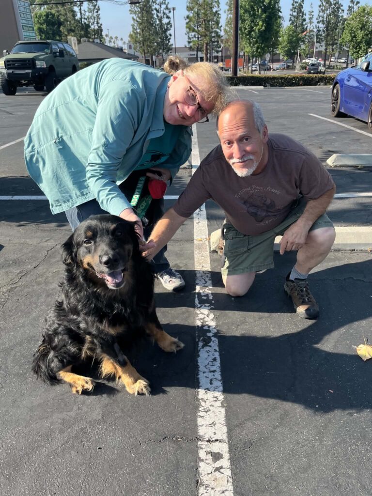 Wasabi sitting happily in a parking lot with his adoptive couple, who are kneeling beside him.