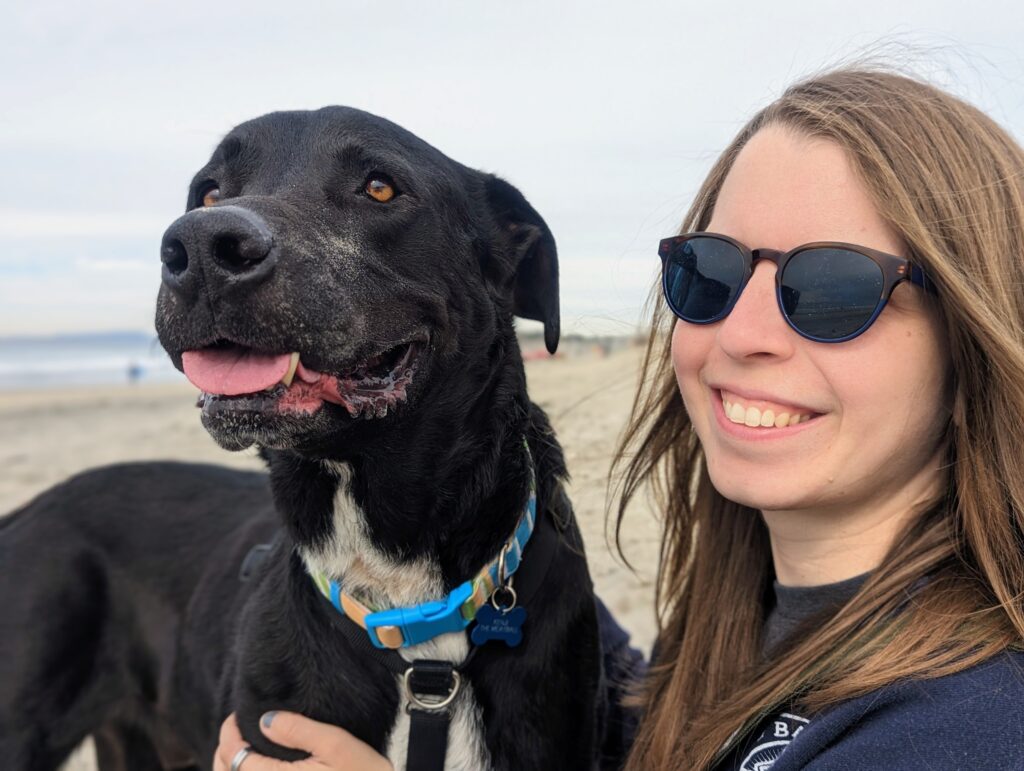Amy, a Labradors and Friends volunteer, smiling beside Kenji, her beloved black Lab hospice foster, during a beach outing.